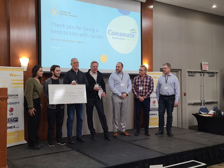 People on a stage exchanging a large novelty sized cheque during a presentation. Coinamatic's logo is present on the overhead projection.
